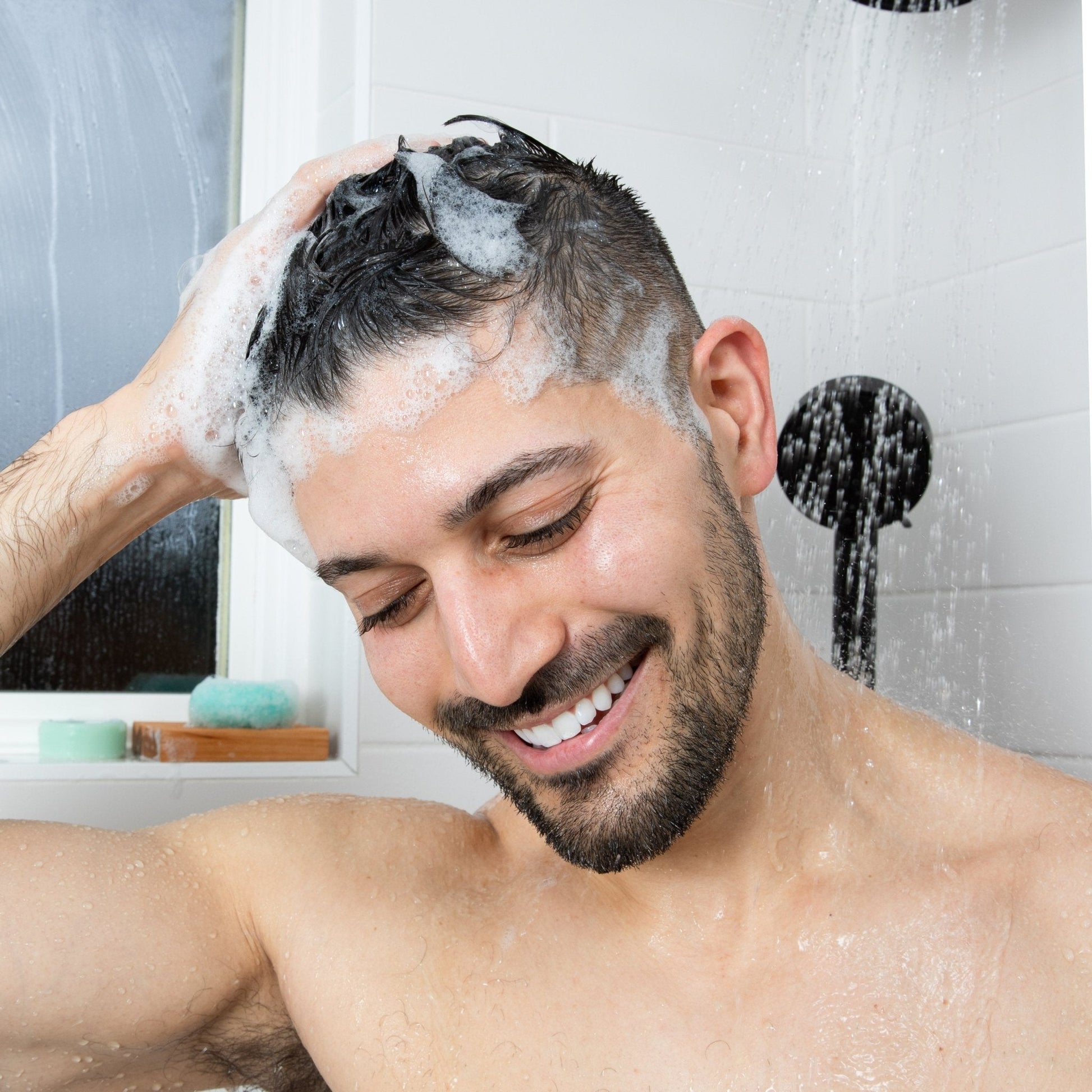 A smiling man washes his hair with a natural shampoo bar by Community Goods.