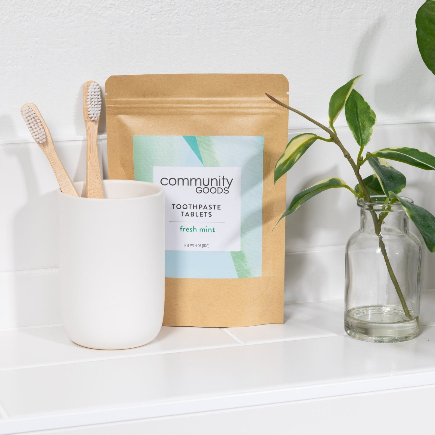 A Kraft paper pouch of the best toothpaste tablets, by Community Goods, is shown on a modern bathroom counter next to two bamboo toothbrushes.