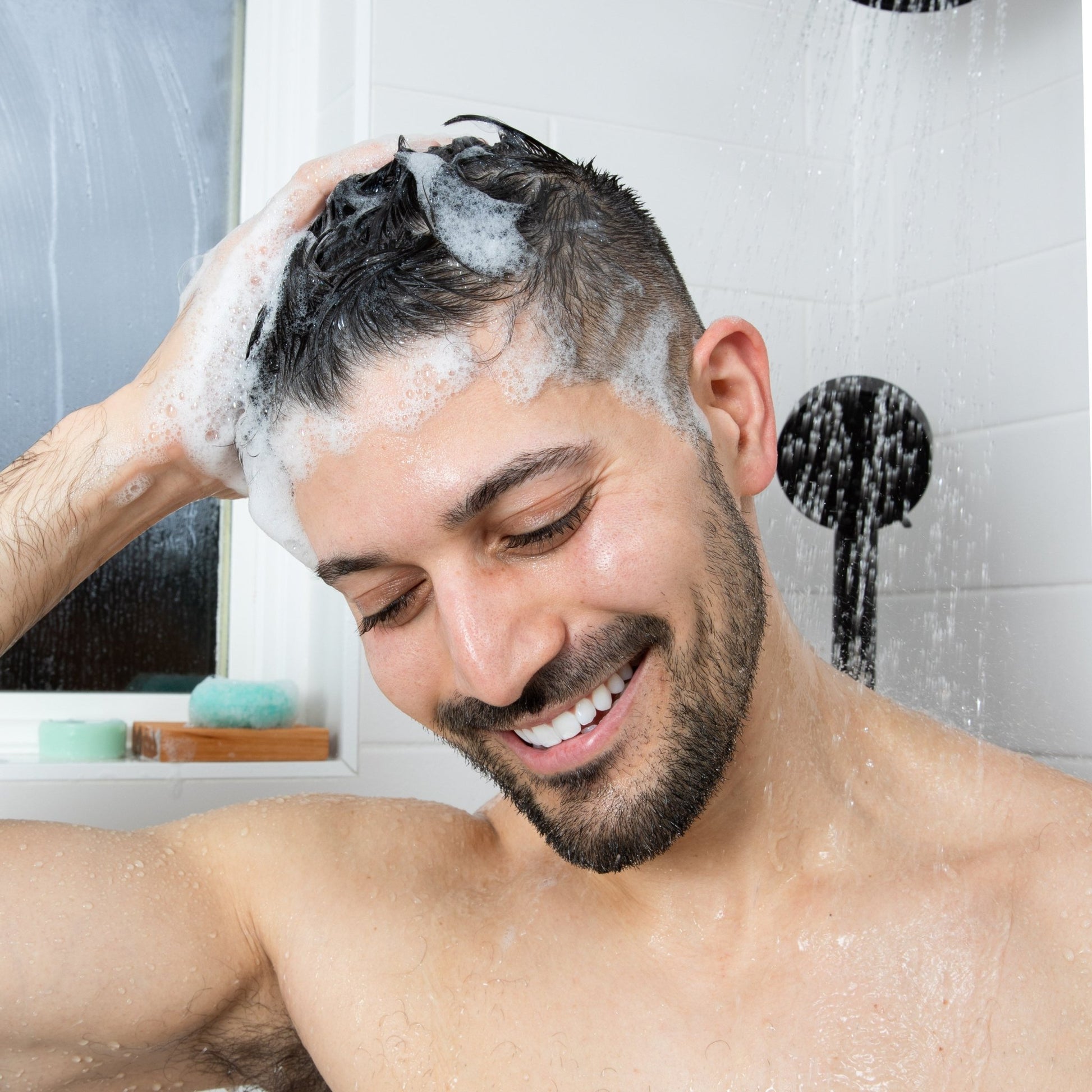 A smiling man washes his hair with a natural shampoo bar by Community Goods.