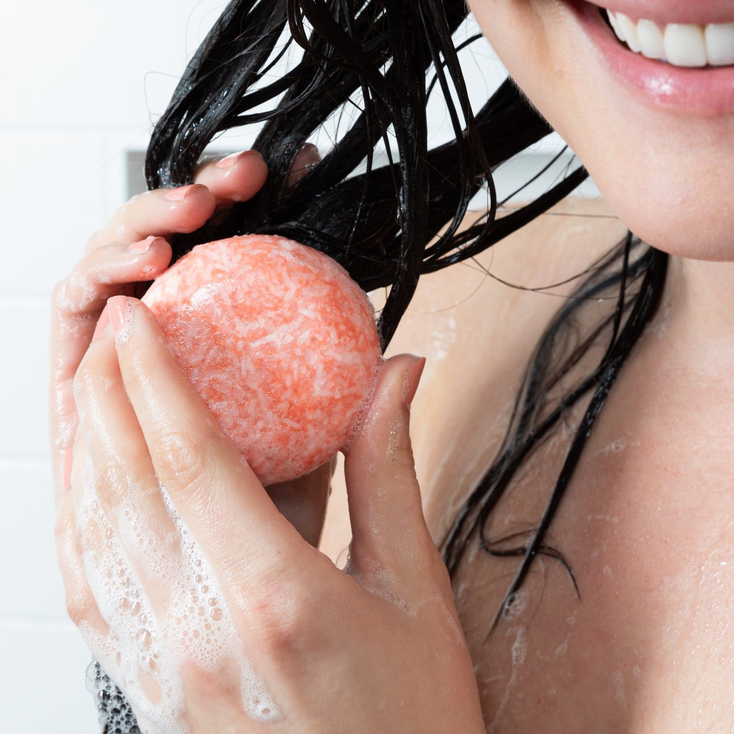 A smiling woman shampoos her hair with a Citrus Squeeze Shampoo Bar by Community Goods.