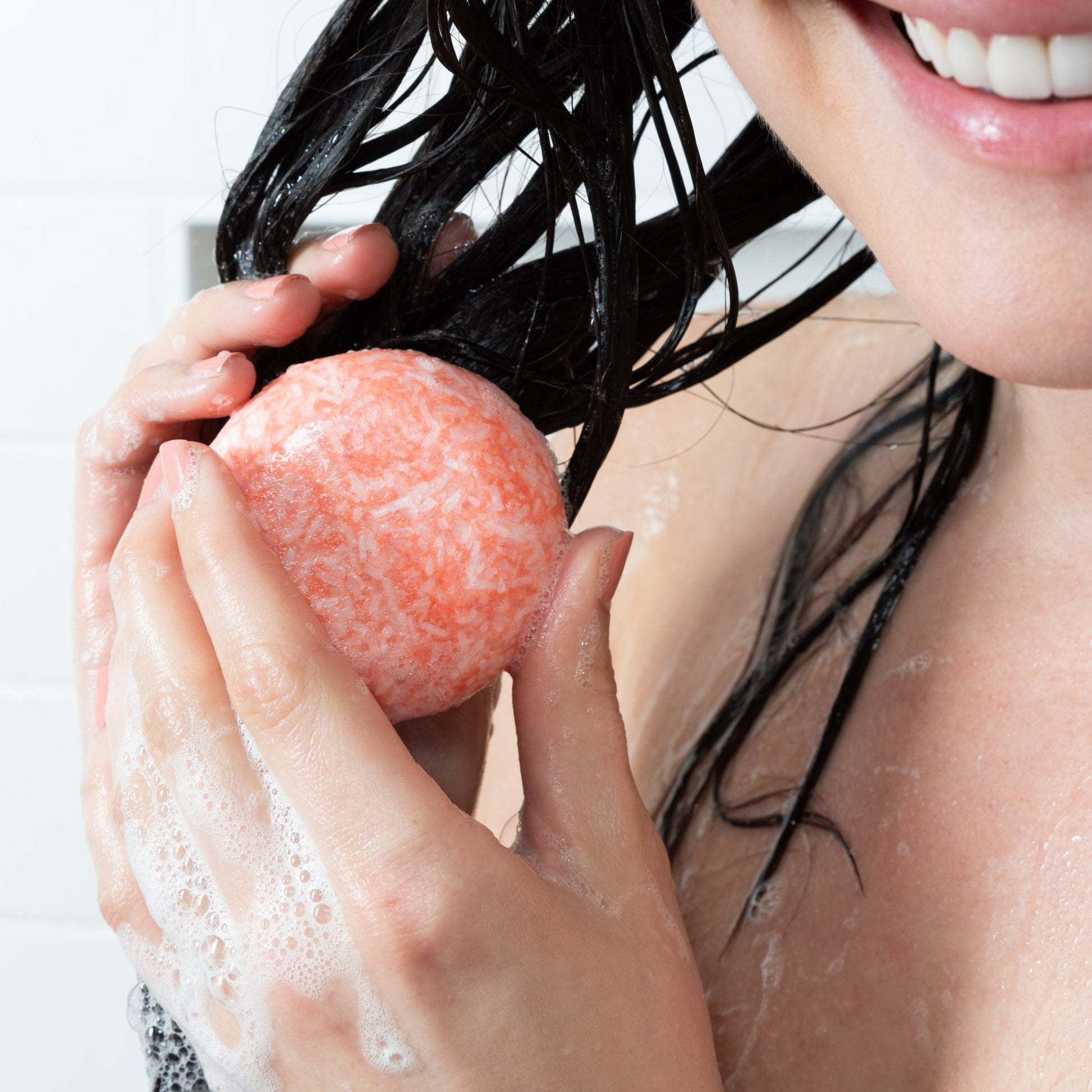 A smiling woman shampoos her hair with a Citrus Squeeze Shampoo Bar by Community Goods.
