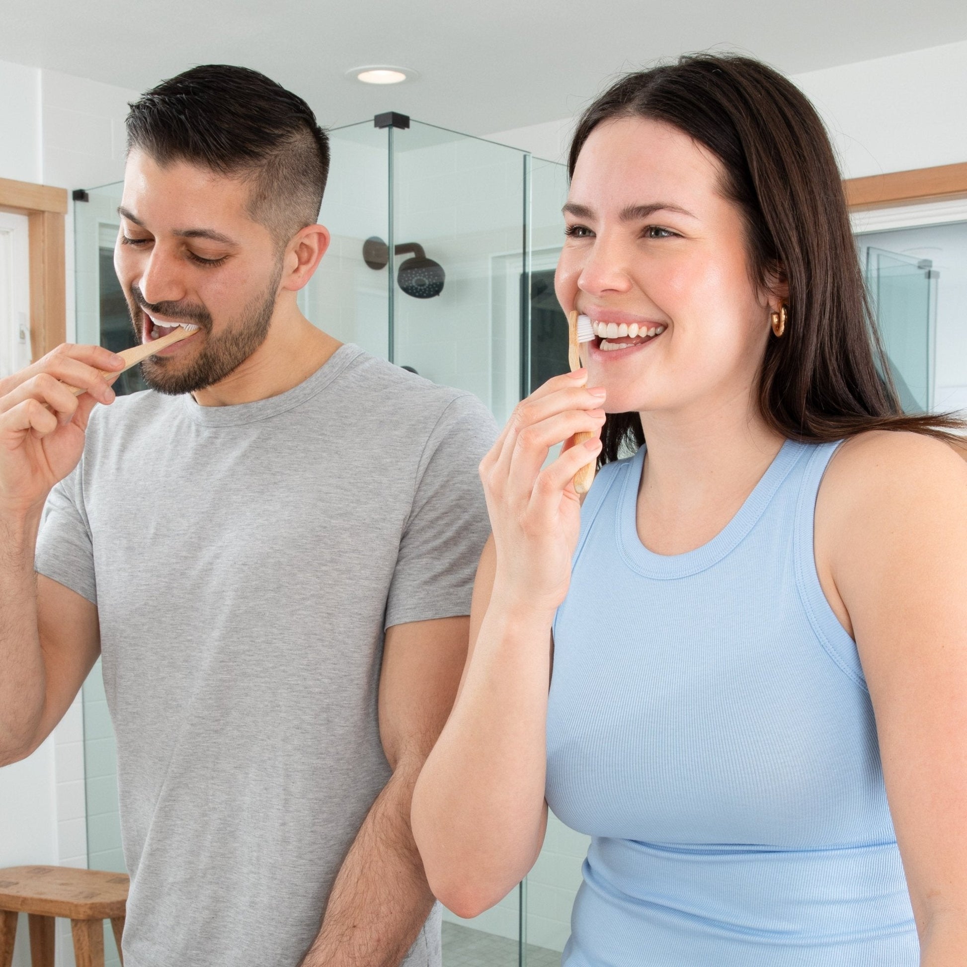 A couple smile while they brush their teeth with the best toothpaste tablets and natural bamboo toothbrushes in a modern bathroom 