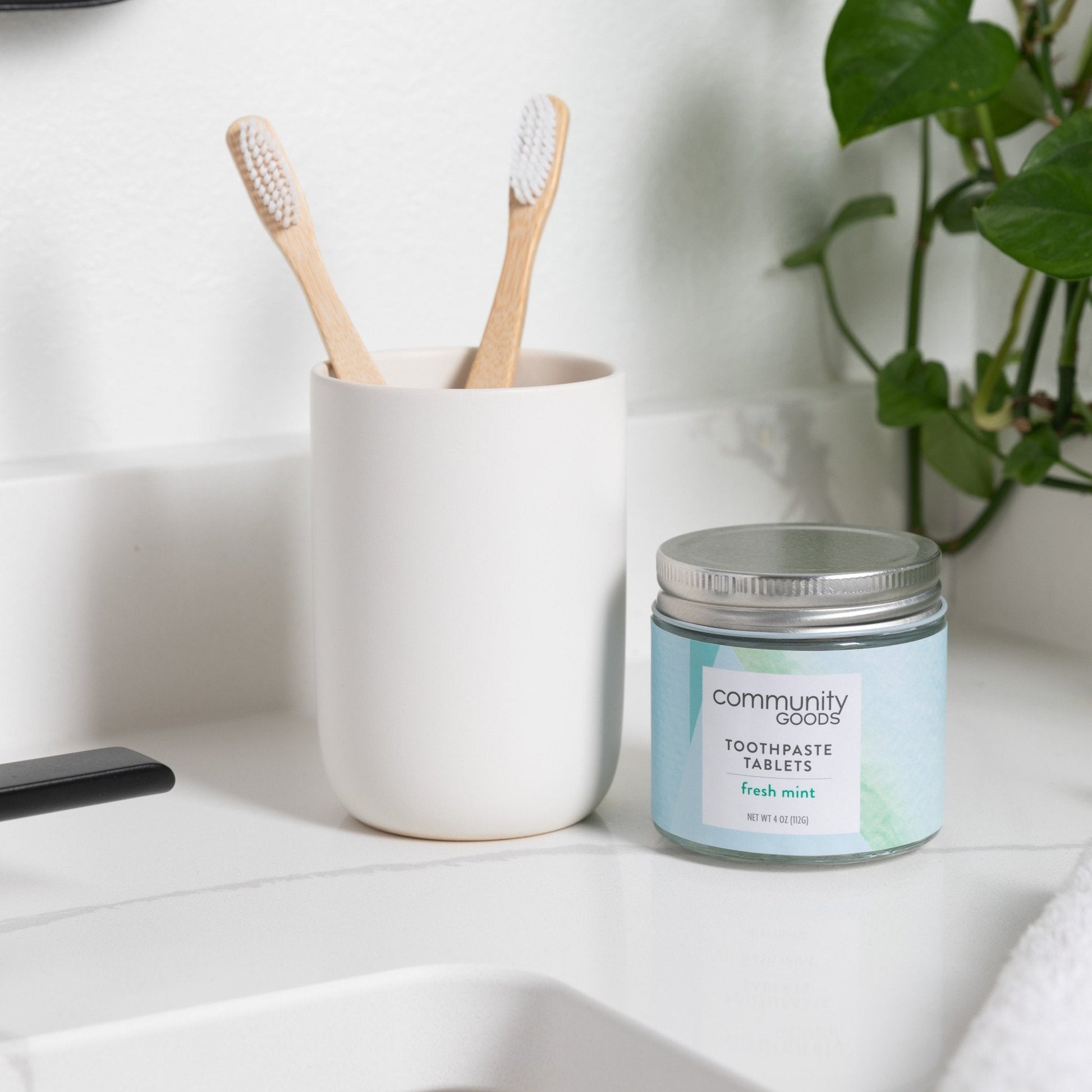 Two natural bamboo toothbrushes, by Community Goods, are shown on a modern bathroom counter next to a jar of the best toothpaste tablets.  